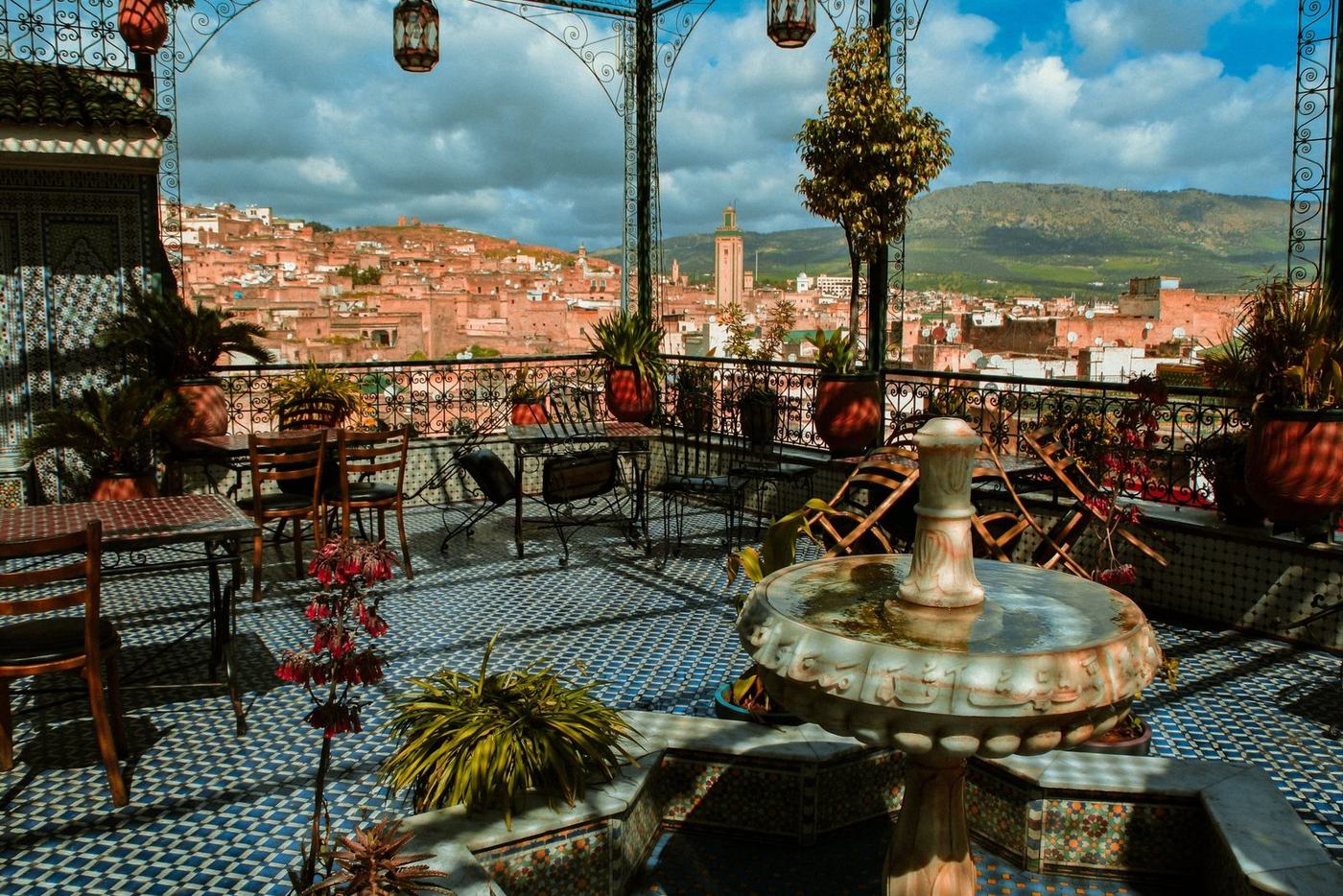 A riad rooftop terrace in Fes, with zellige tilework and a fountain, overlooking the medina and the hills beyond